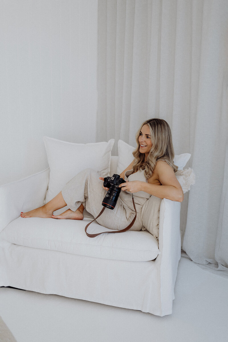 a blonde woman sitting on a sofa wearing a cream suit and holding her camera while she's photographed for her branding photographs