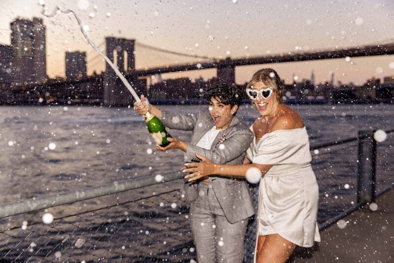 A couple popping a bottle of champagne by the water.