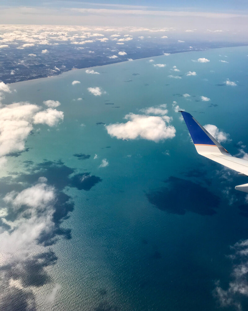 a picture out of a plane window flying over a body of water, white clouds, a portion of the plane wing, and the shore line can be seen