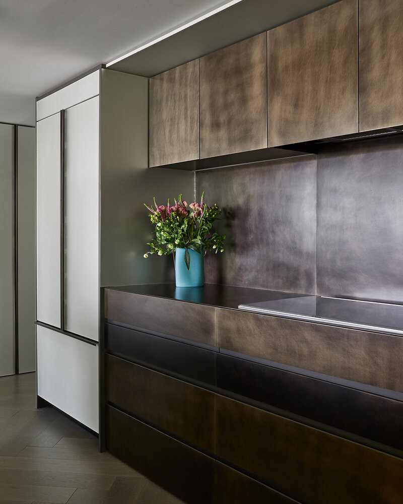 A before and after transformation of an outdated kitchen design to a modern, minimalist design featuring bronze cabinetry and a vase of vibrant flowers