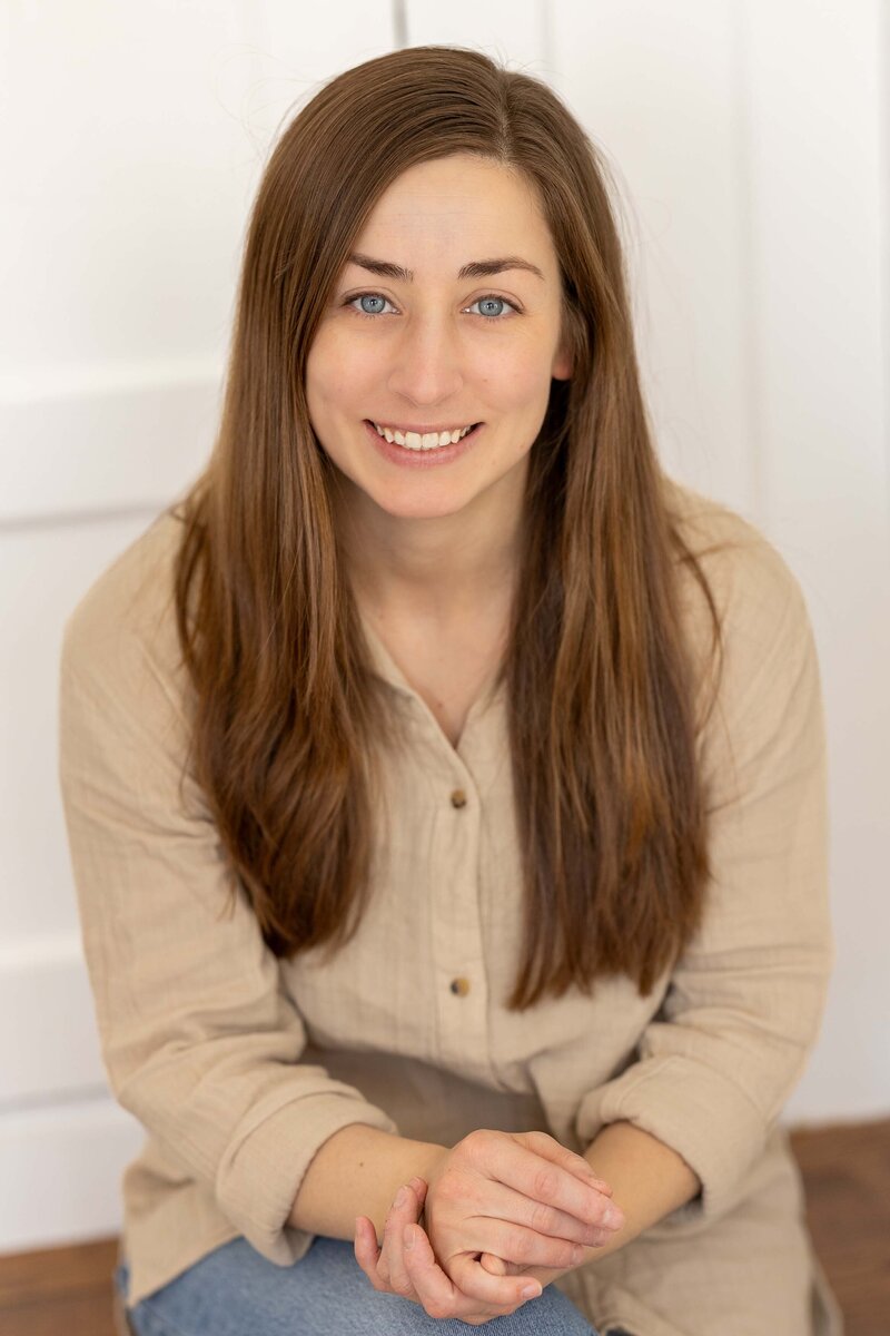 Portrait of Alicia Seeley, a professional New Jersey family photographer, smiling and sitting indoors in a button-down shirt
