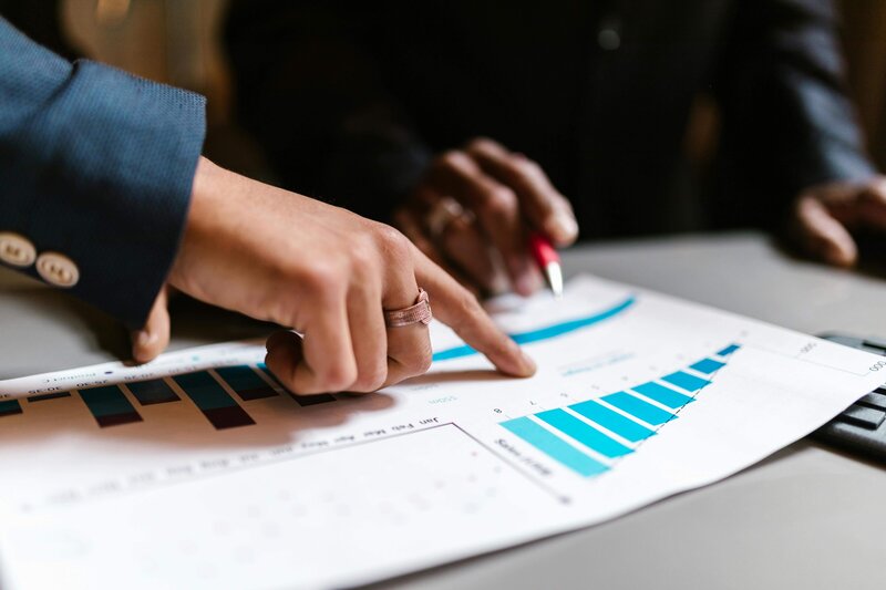 Close-up of two hands pointing at a printed graph on a paper document. One person holds a pen while the other points at a specific bar on the chart, suggesting collaboration and analysis.