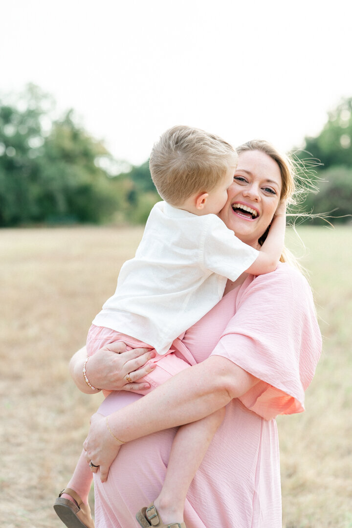 A pregnant mother holds her son while he kisses her on the cheek. The mother is smiling in this candid shot captured by an Austin Maternity Photographer
