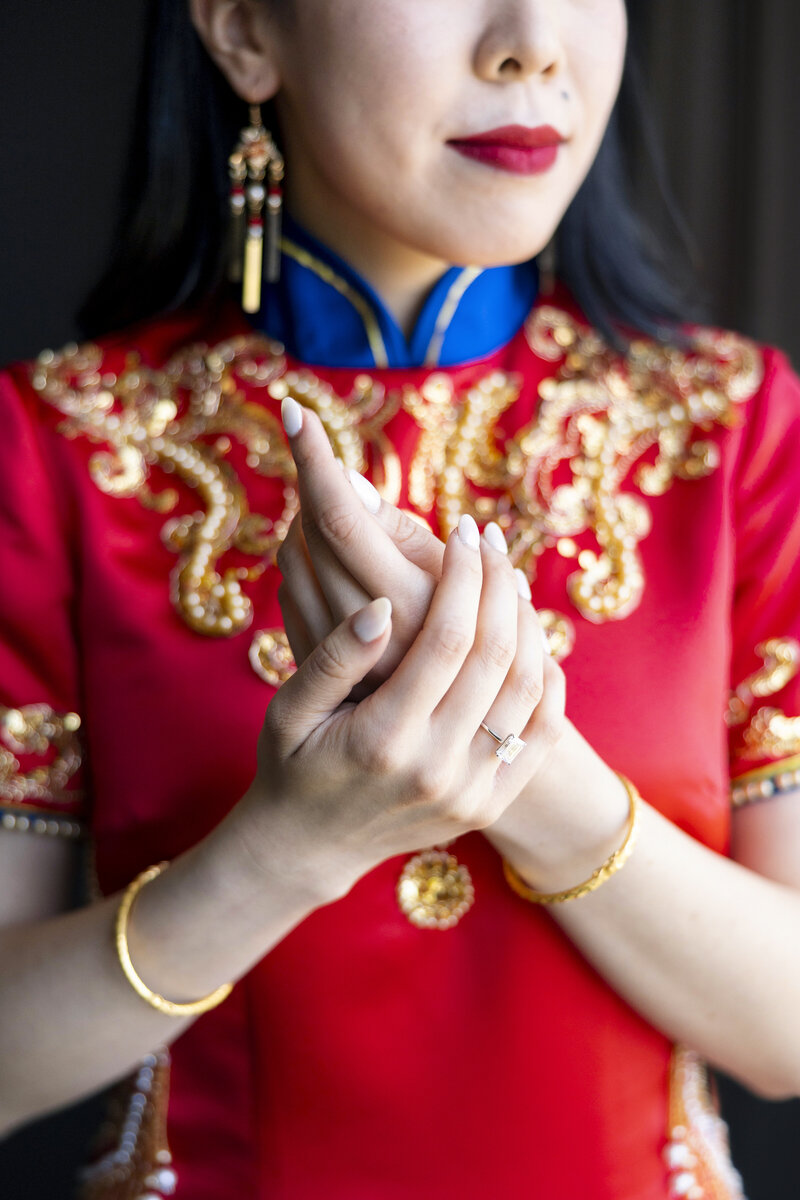 A woman in a red and gold embroidered dress, with elegant jewelry, clasps her hands together. She wears gold bangles and drop earrings, and her nails are painted white. Her face is partially visible, focusing on her lips and chin.