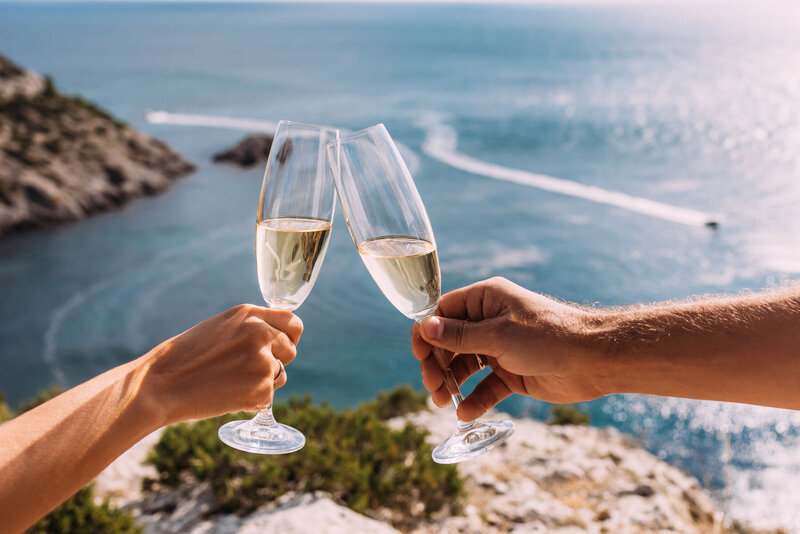 Two people clinking champagne glasses with an ocean view and rocky coastline in the background.