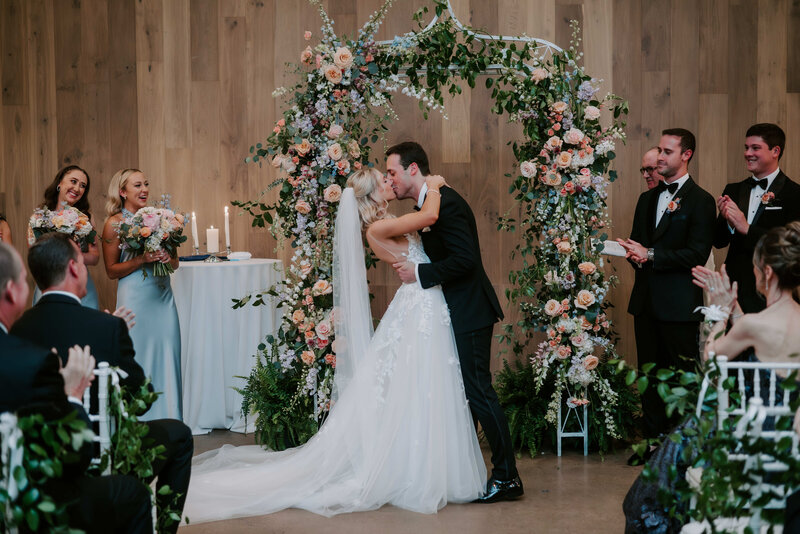 A romantic indoor wedding ceremony with a bride and groom sharing their first kiss under a floral arch decorated with roses, greenery, and pastel blooms, surrounded by bridesmaids, groomsmen, and guests applauding.