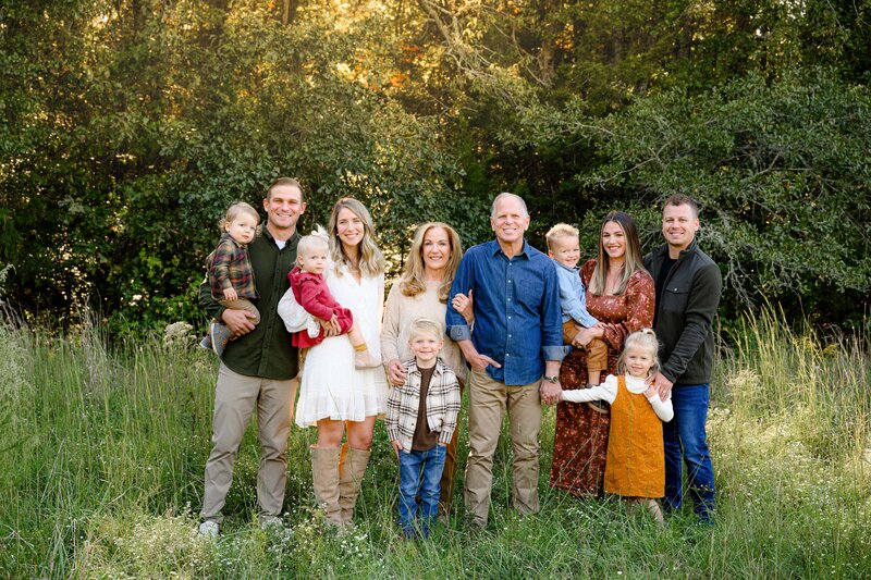 A family of eleven, including five children, stands together in a grassy field with trees in the background. They are smiling and dressed in casual clothing.