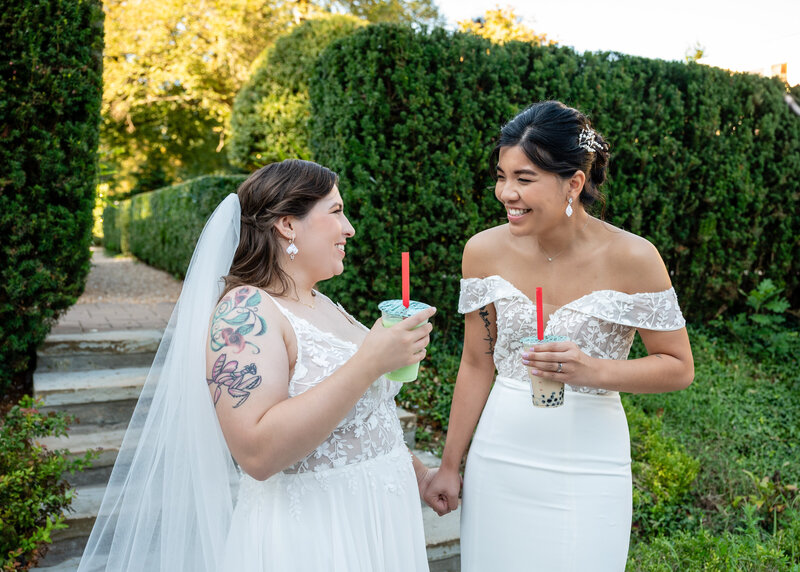 laotian bride in off the shoulder white gown with a curvy white bride in a spaghetti-strap gown holding hands, smiling at one another, and drinking boba tea while walking on a stone path in a green hedge garden