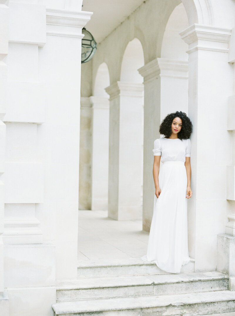 woman with short black curly hair in white dress leaning against a pillar