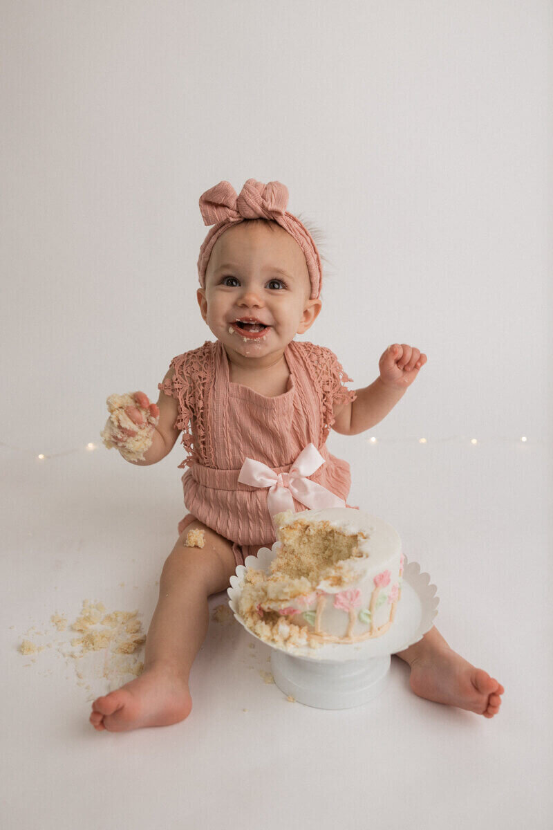 A baby girl with a pink headband and romper smiles while holding cake in her hand. A half-eaten cake decorated with pink flowers sits on a white stand in front of her, with cake crumbs scattered around.