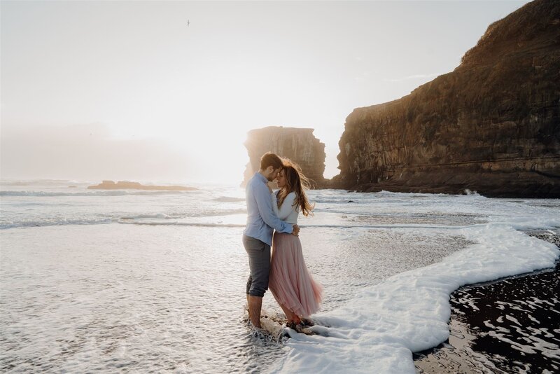 A couple wearing a blue shirt and a pink skirt standing in the water during an engagement photoshoot in Muriwai, Auckland, photographed by waikato photographer haley adele photography