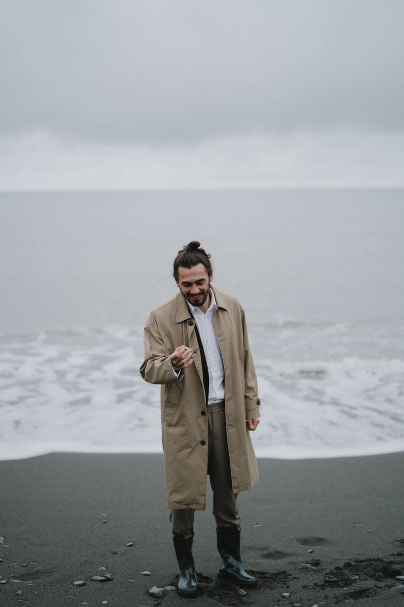 Ein Mann im Trenchcoat und Gummistiefeln lächelnd am Strand. Hinter ihm die Gischt – bereit für Abenteuer und positive Veränderungen