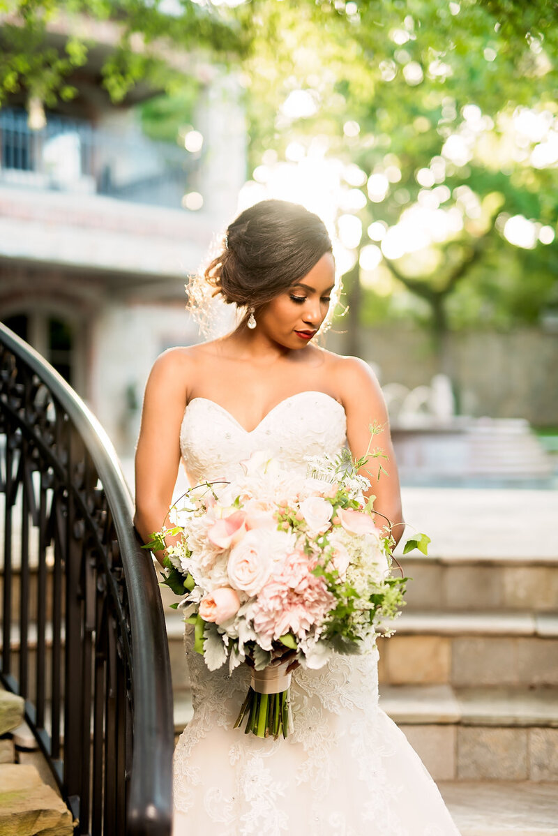 A bride looks down on an outdoor staircase, holding a large pink and white bouquet.