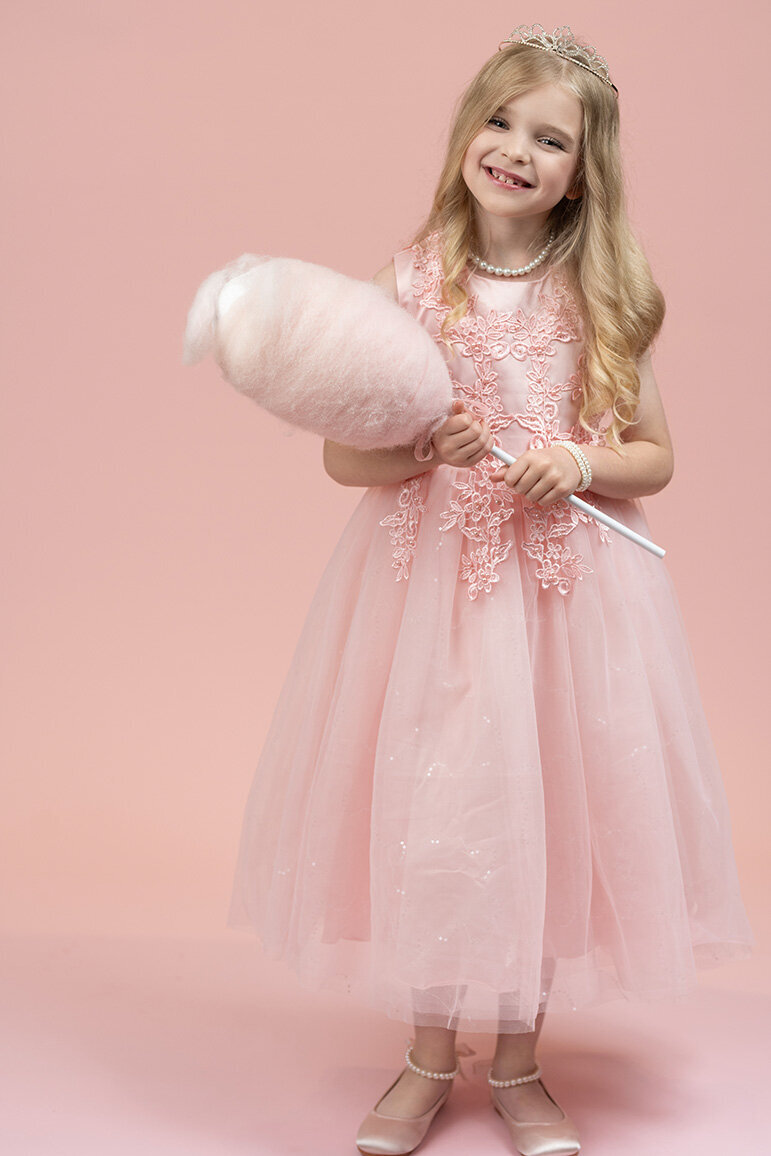 A young girl in a pink dress and tiara smiles while holding cotton candy. She conveys joy and innocence against a matching pink background.