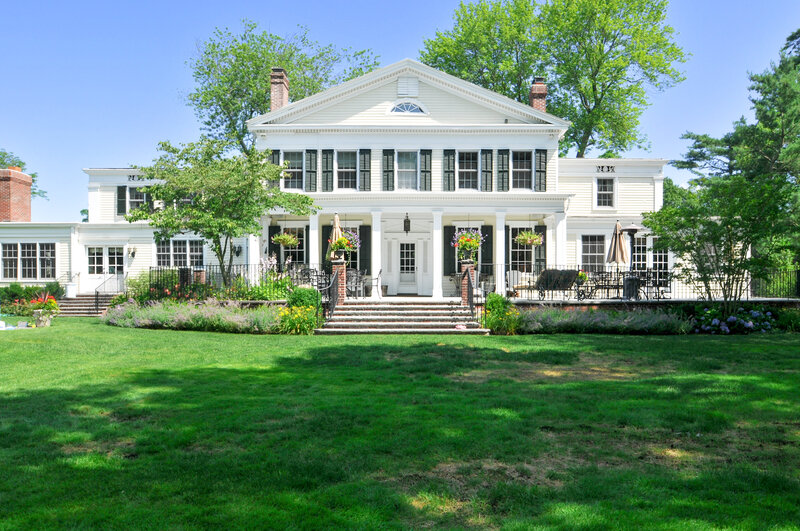 Renovated Colonial estate in Brookville featuring white clapboard siding, black shutters, and a landscaped backyard.