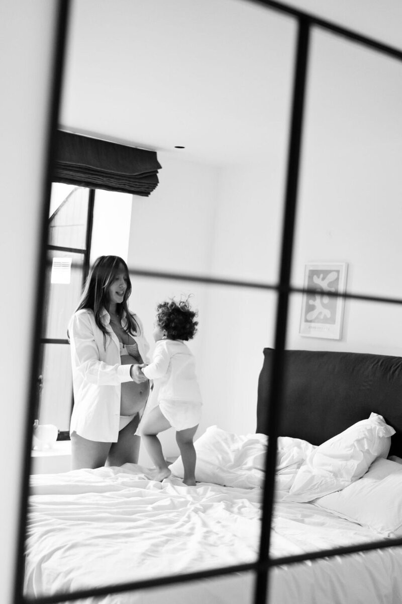Toddler holding hands with his pregnant mother as he jumps on her bed during a maternity photo shoot with Jess Morgan Photography