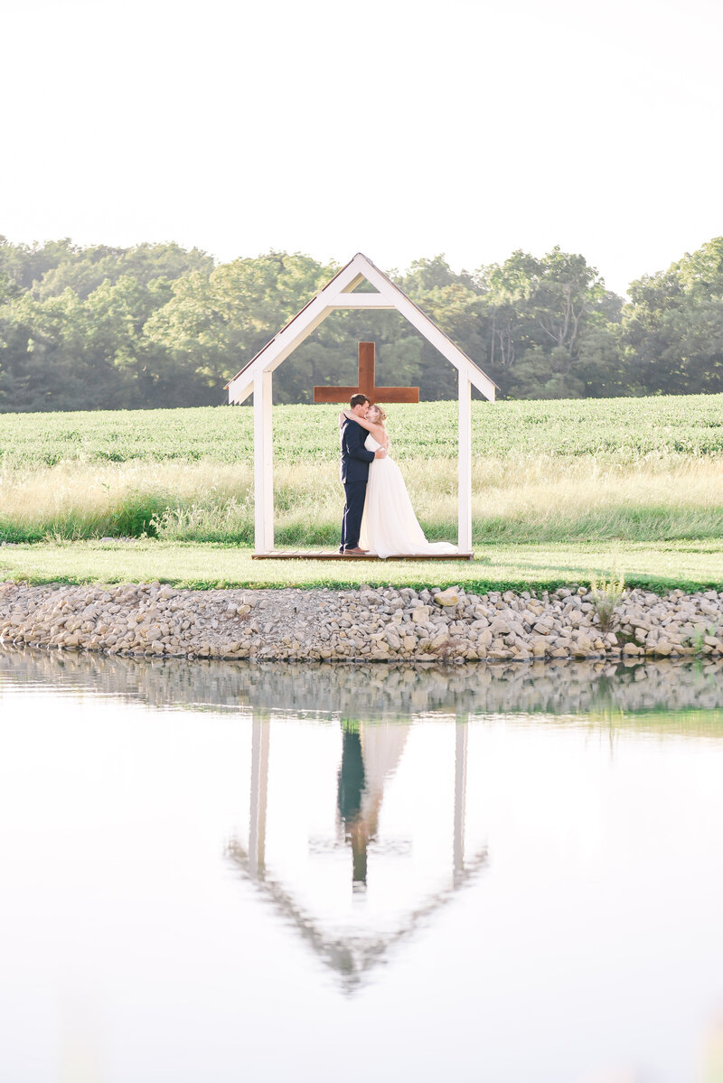 A bride and groom are reflected on the lake as they pose in front of a cross arbor.