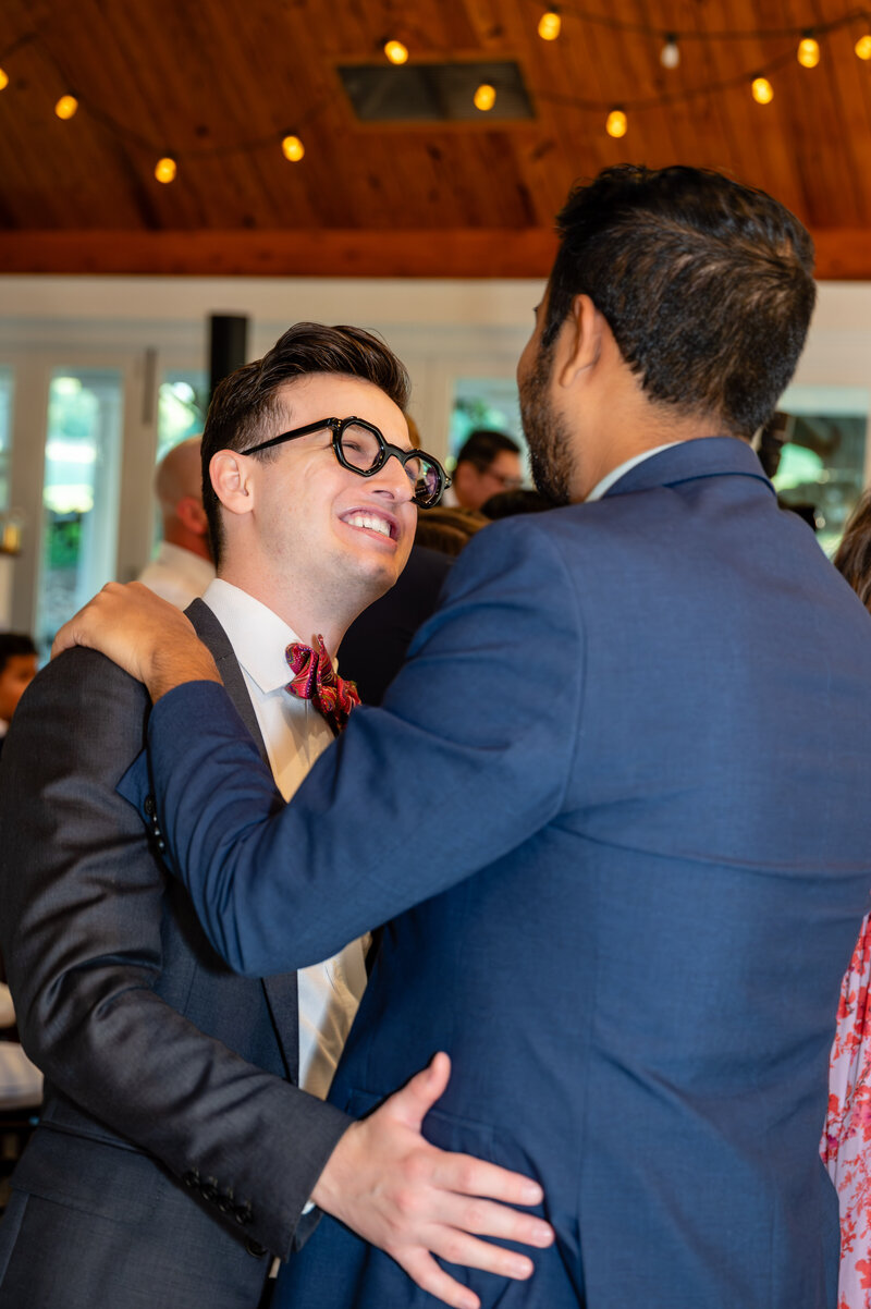 gay white man in a black tux with red bow tie and black rimmed glasses embracing a gay indian man in a blue suit dancing on the dance floor