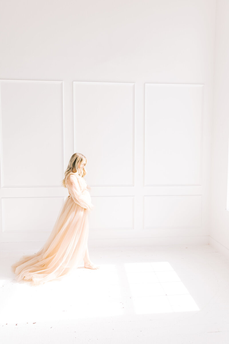 A mom to be walks through an open room holding her bump in a pink maternity gown