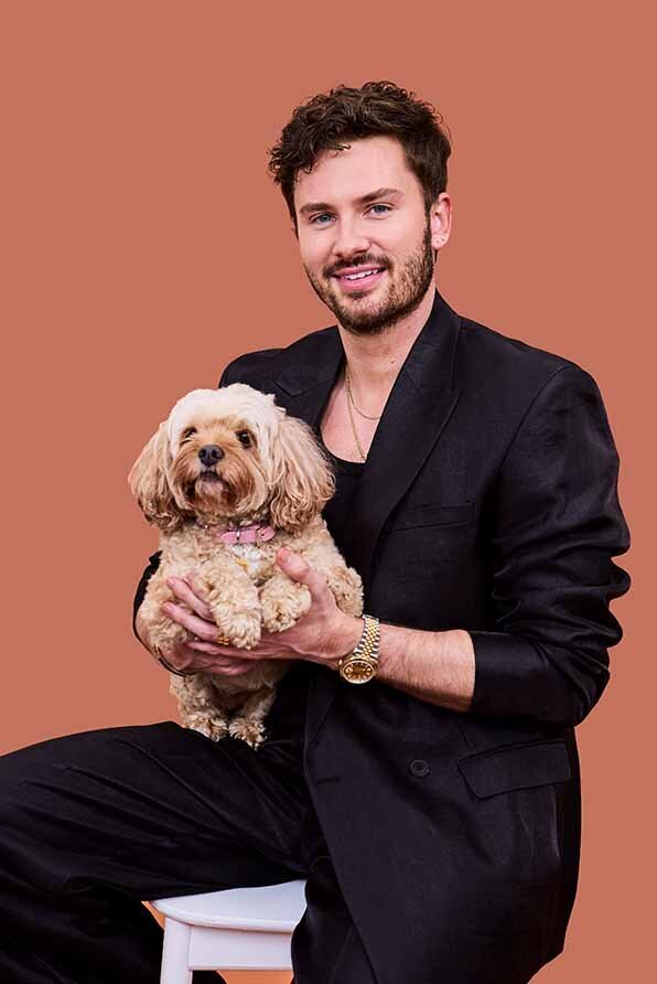 Male Hairdresser, Jordan Hone, dressed in a sleek black suit holding a small fluffy dog with a pink collar against a warm-toned backdrop.