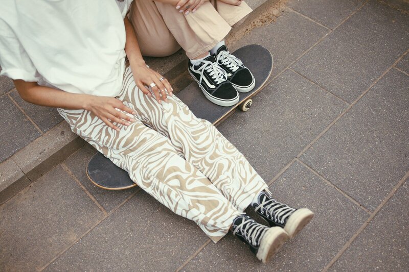 two people sitting on a curb resting their legs on a skateboard