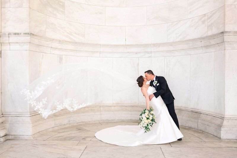 A groom in a black suit and bride in a white gown kiss passionately. The brides veil flows dramatically in the air, and she holds a bouquet of white flowers. They stand in front of an elegant, curved marble wall.
