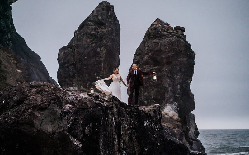 a couple in wedding attire explore rocky, sea stacks as the sun sets. They explore by lantern light 