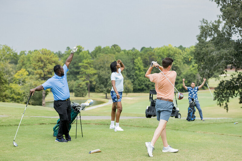 Group of golfers celebrating on the green at Woodcreek Country Club in Columbia, SC. A man in a peach shirt finishes his swing while others react with excitement. The lush golf course, lined with trees and rolling hills, creates a scenic backdrop. Small business branding photography in Columbia, SC, highlighting golf lifestyle, recreation, and community.