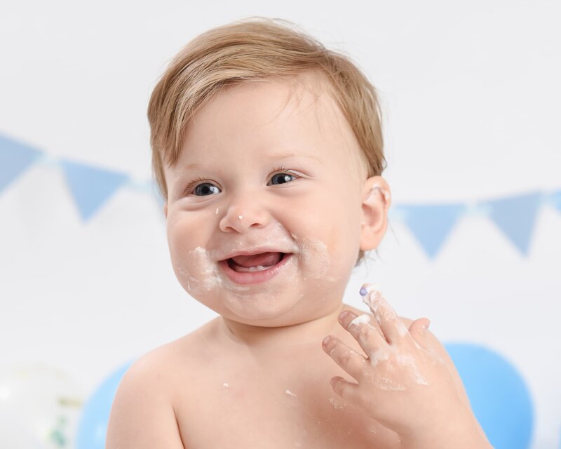 A baby boy with blonde hair and frosting on his face, smiling cheerfully during his cake smash photoshoot, with soft blue bunting in the background