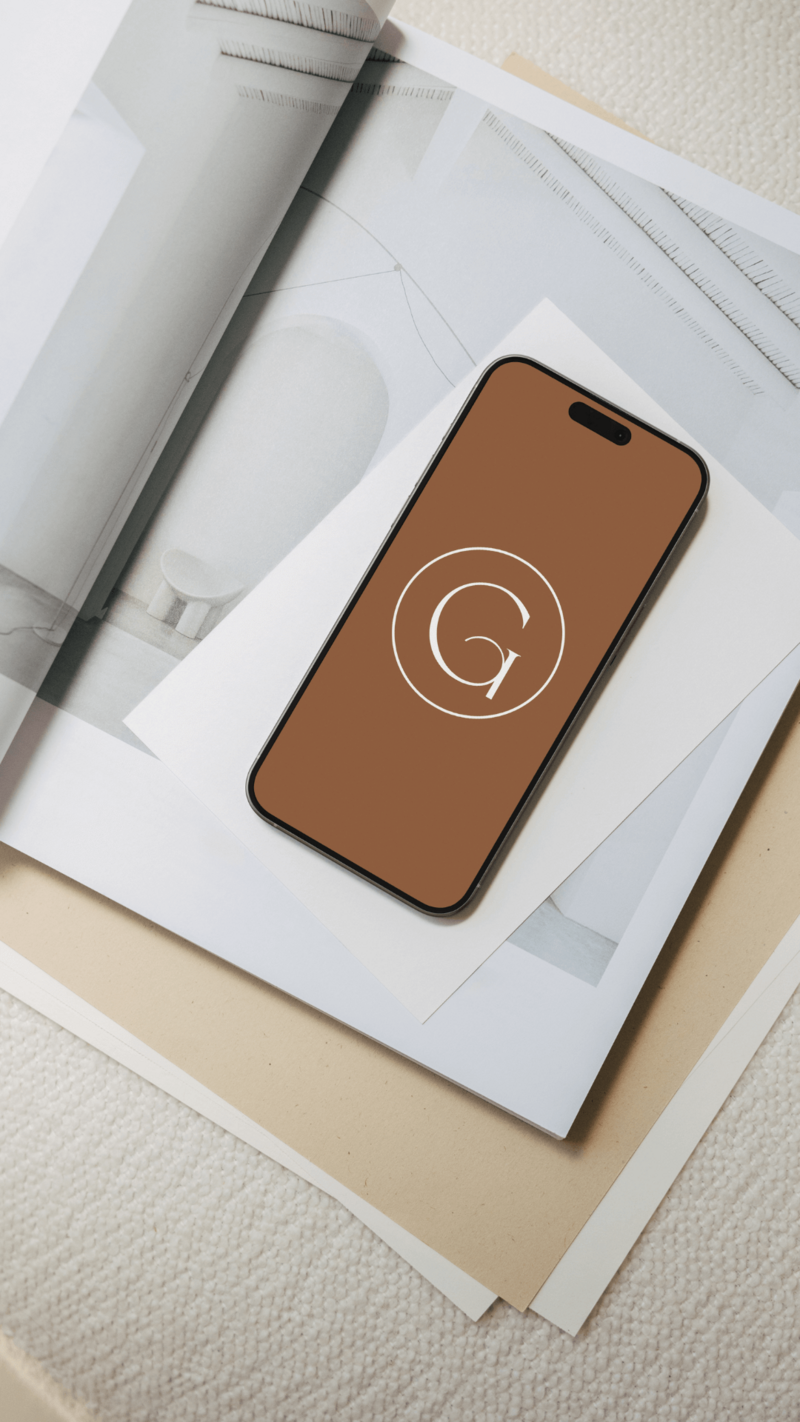 A smartphone with a brown screen displaying a white letter G in a circle rests on layered papers and an open magazine, capturing the inspiration behind modern hair salon website design on a textured beige surface.