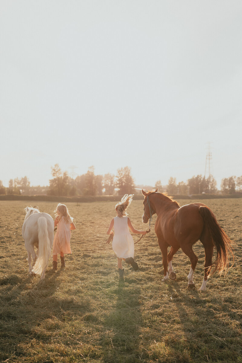 Photo of 2 young girls running through a farm field holding onto their horses who are running beside them, taken by a langley photographer.
