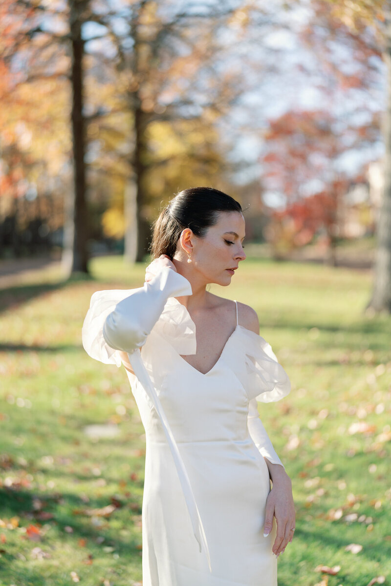 Bride in an elegant off-shoulder gown stands in a sunlit park with vibrant autumn foliage, softly touching her hair in a moment of quiet grace.