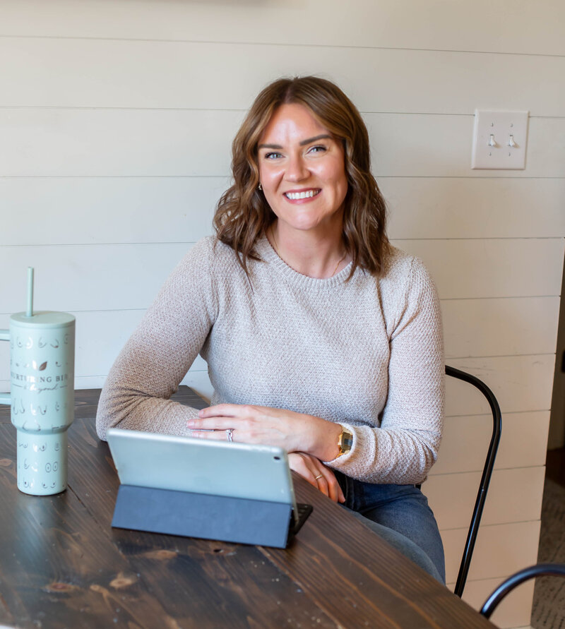 Certified Breastfeeding consultant, Sarah, sitting at a wooden desk with an iPad smiling at the camera wearing a cream colored sweater