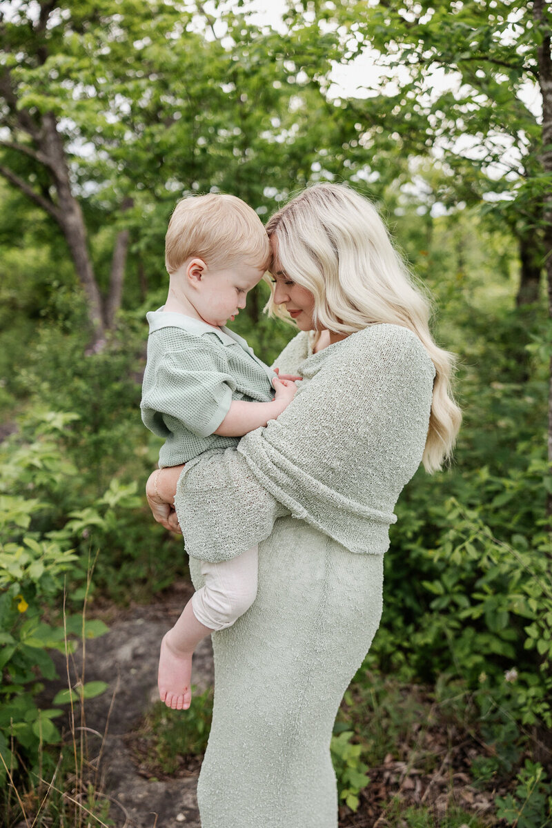 Maternity portrait of expecting mother carrying toddler on her belly in the woods.