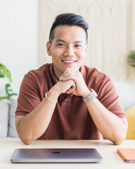 Sho Dewan, young Asian man, founder of Workhap, wearing a short-sleeved brown top, sitting at a desk with a closed laptop in front of him