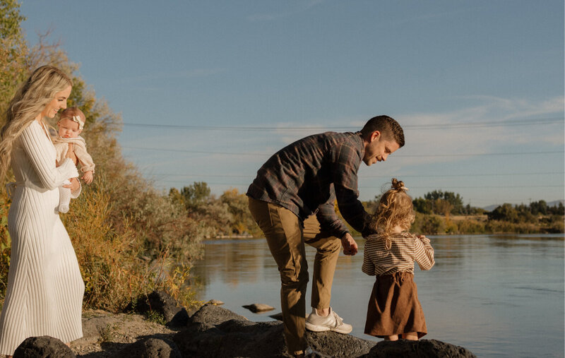 A dad is holding the hand of his toddler and the edge of a lake. The wife is behind, holding their baby.