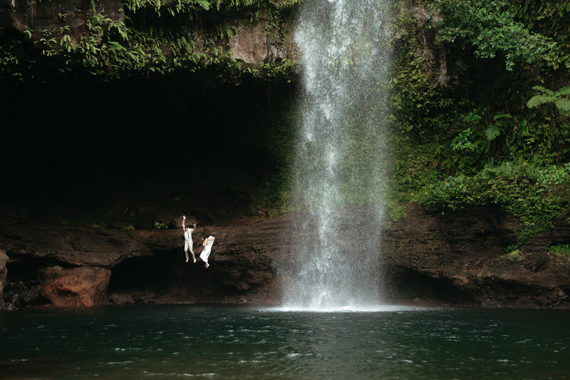 Unique Elopement Ideas | Eloping couple in mid air after jumping off into the water, a huge waterfall is right next to them