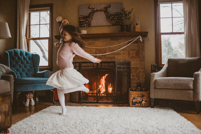 young girls twirl in front of the fireplace while sister watches, smiling, hiding under living room chair