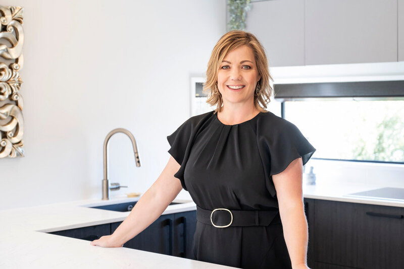 Anna James, a professional real estate agent with blonde hair, smiles warmly while standing in a sleek kitchen wearing a black belted dress, showcasing confidence and approachability