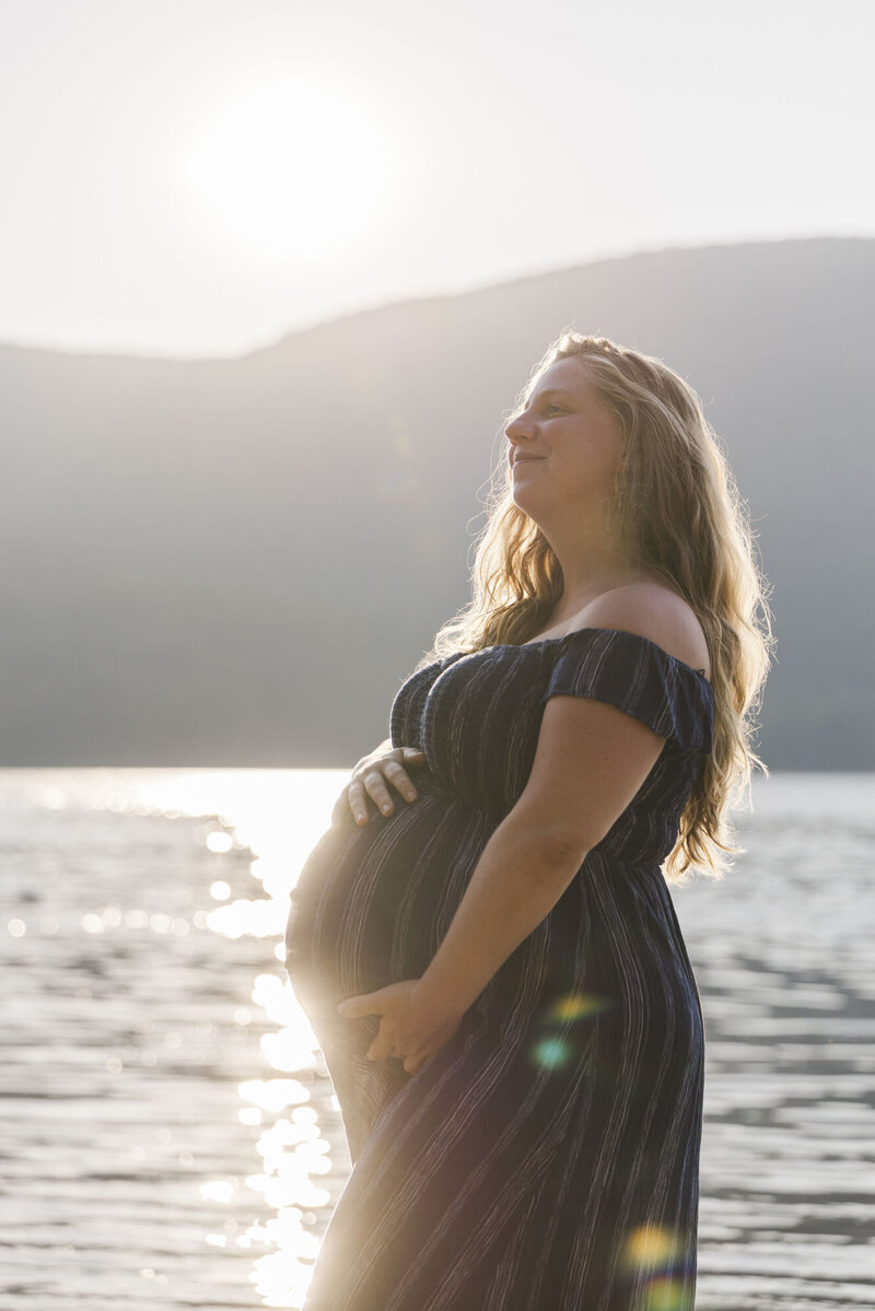 Expectant mother holding her belly by the Hudson River at sunset during a Hudson Valley maternity session