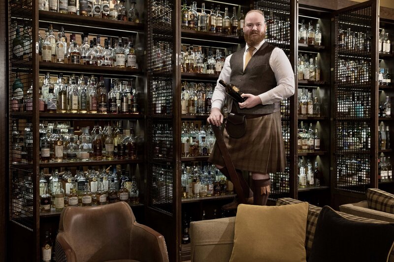 Guy in a kilt holding a wine bottle standing on step stool with wine cabinets behind him