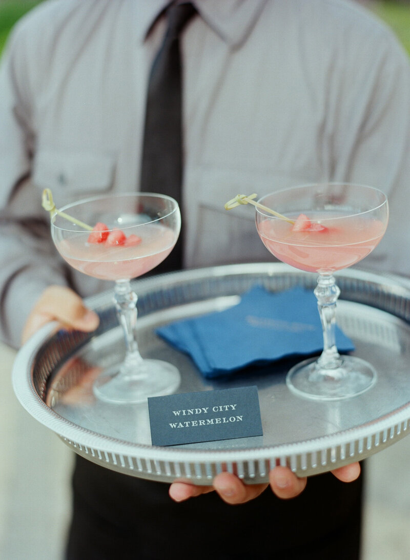 Server holding a tray with two elegant cocktails labeled 'Wind City Watermelon,' perfect for a wedding reception.