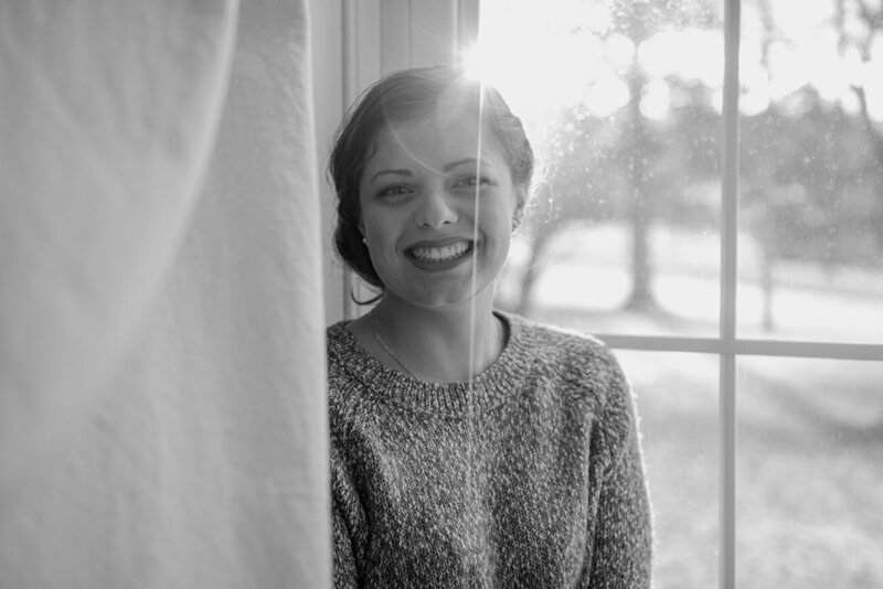Photo shows a girl smiling at the camera in black and white. She's wearing a sweater and sitting in front of a window. There's some sunlight flaring through the window.