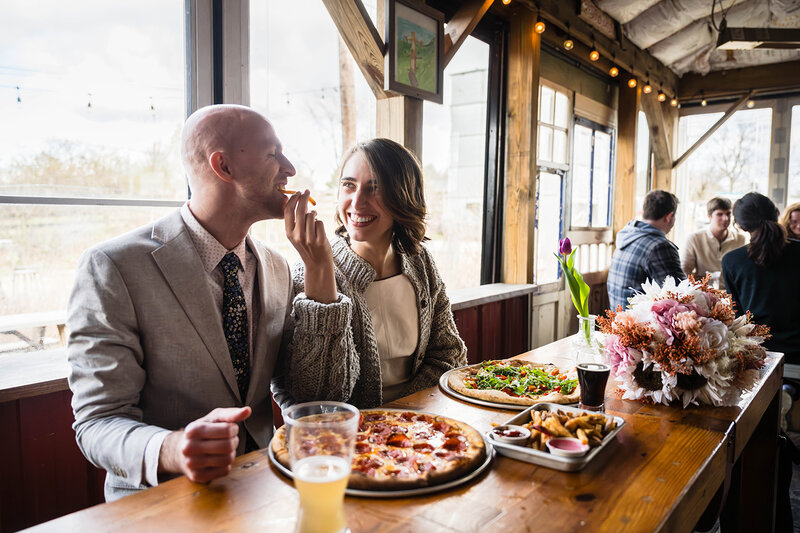 A couple on their elopement day sit a long table at Rising Silo Brewery in Blacksburg, Virginia. At their table are two beers, two medium pizzas with various toppings, and a small tray of fries with two sauces. One newlywed smiles as she feeds a fry to her partner who playfully takes a bite.