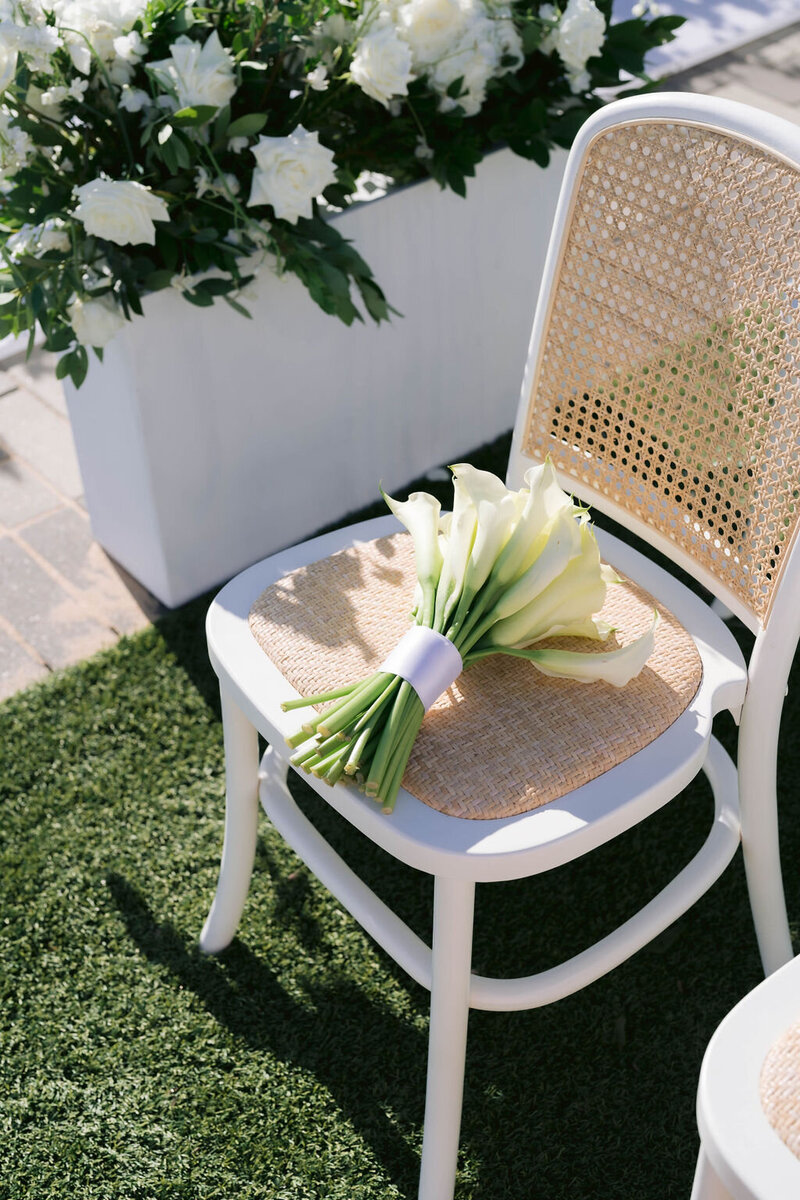 Calla lily bouquet resting on a white cane-back chair next to a white floral planter at an outdoor Houston ceremony.