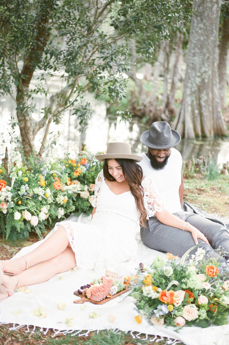 married couple on a picnic blanket with charcuterie bride wearing a short lace dress and a hat groom wearing a white shirt and gray suit and hat with spring colored flowers
