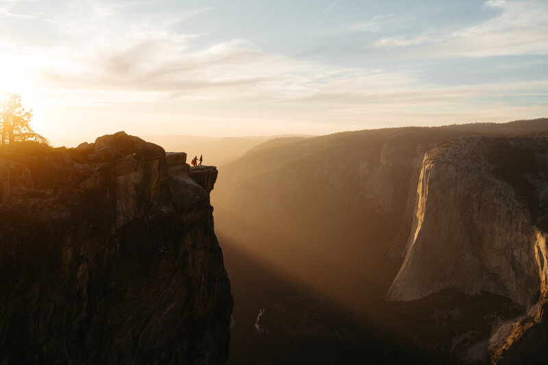 Adventure Elopement Ideas | Couple stands on a huge mountain cliff at Taft Point with a golden sunset behind them shining onto the mountains 