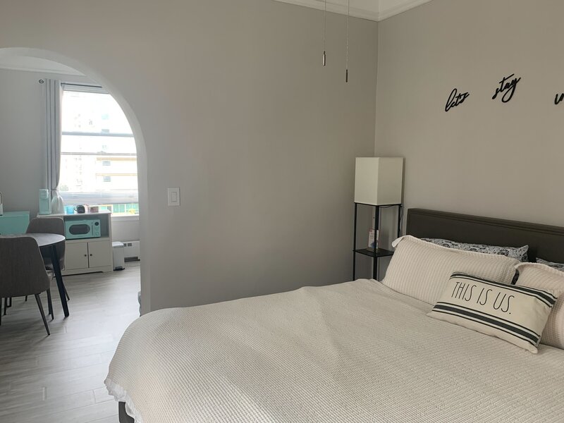 Picture of a bedroom with a king bed done in white, a lamp, and the words "let's stay" on the wall. You can see through a doorway to the next room which has a table, two chairs, a mint green mini-fridge and a mint-green microwave. The next room also has a window full of light.