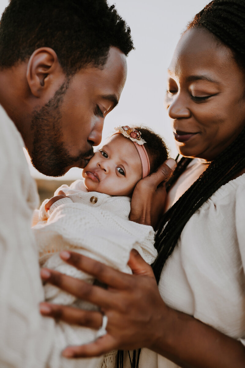 A dad kissing his newborn daughter on the cheek as mom looks on smiling at Murrell Park in Flower Mound, Texas
