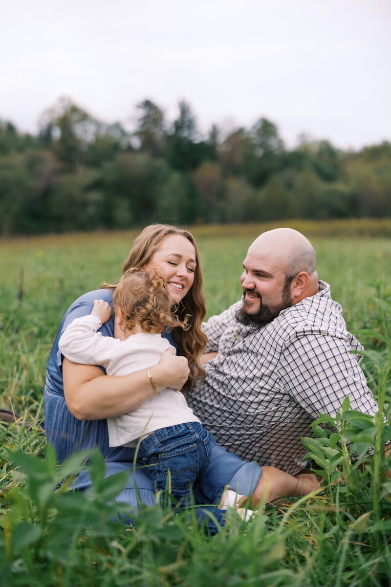 Courtney Paige with her husband and son sitting together in a field, smiling during a family photo session.